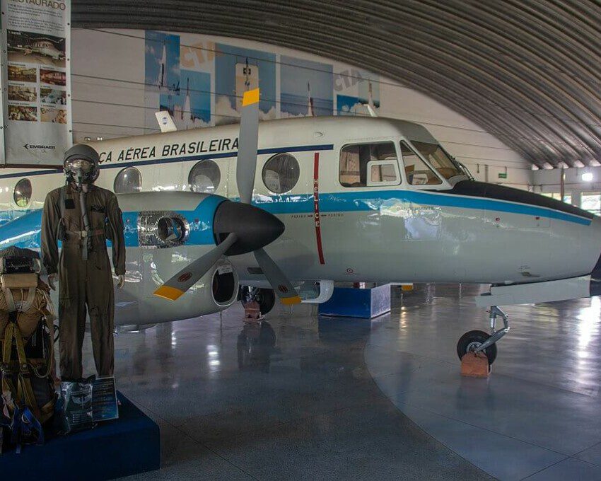 Foto que ilustra matéria sobre o Museu Aeroespacial Brasileiro mostra o interior de um galpão com um pequeno avião. Ao lado dele se encontra um manequim com roupas de aviação e outros equipamentos.
