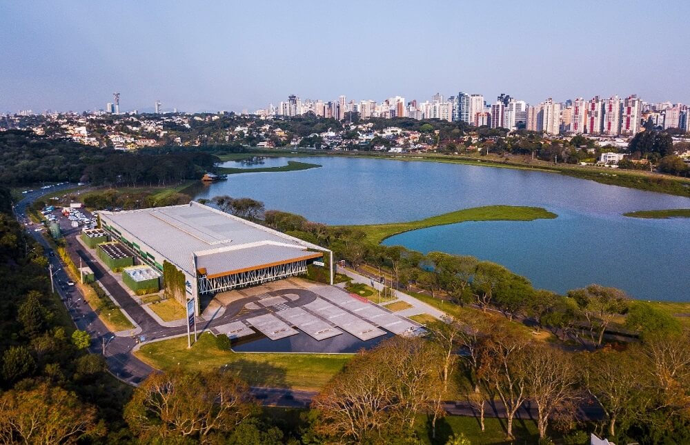 Foto que ilustra matéria sobre o Parque Barigui mostra o Centro de Eventos Positivo visto de cima, um pavilhão localizado ao lado do grande lago do parque.