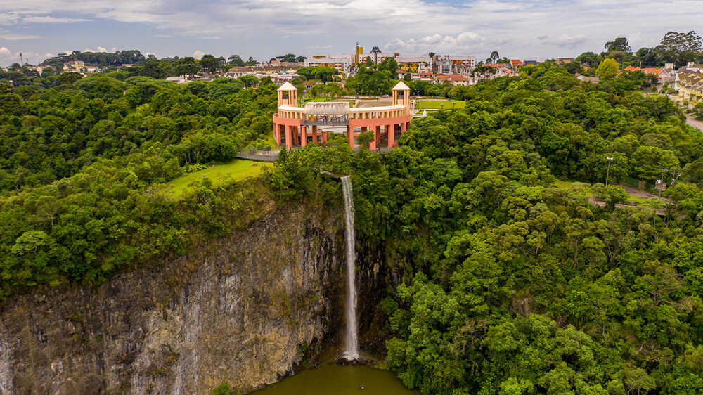 Foto que ilustra matéria sobre o Parque Tanguá mostra uma vista panorâmica da queda d'água que sai do meio de dois grandes paredões de rocha e termina em um lago. Na parte de cima dos paredões fica o mirante do parque.