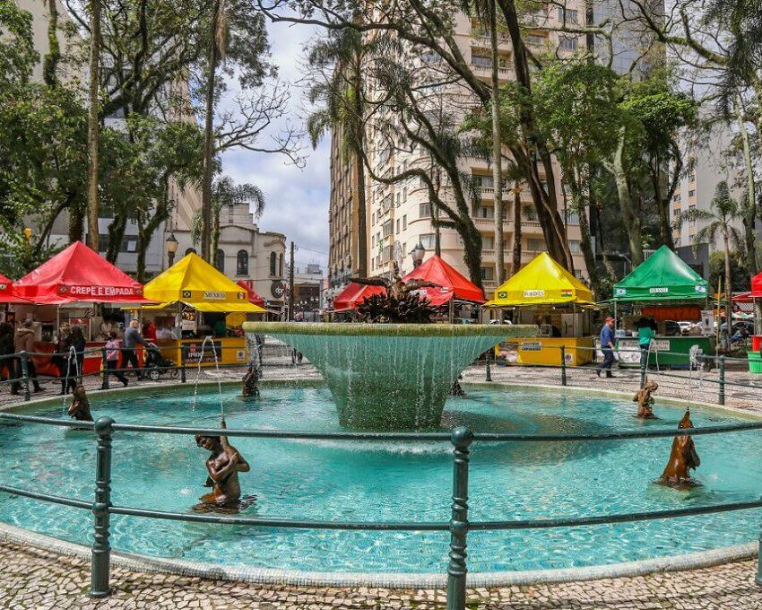 Foto que ilustra matéria sobre a Praça Osório, em Curitiba, mostra o chafariz localizado no centro da praça cercado de barraquinhas de comida
