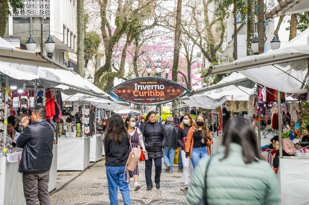 Foto que ilustra matéria sobre a Praça Osório mostra barraquinhas brancas com pessoas circulando no meio na praça, com o letreiro da feira especial Inverno Curitiba ao fundo.