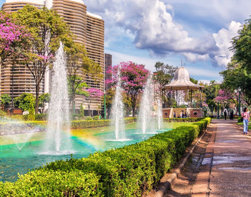 Foto que ilustra matéria sobre a Praça da Liberdade em Belo Horizonte mostra um trecho da praça onde há uma fonte de água com chafarizes, um coreto e o Edifício JK ao fundo.