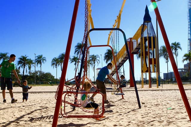 Área coberta por areia no Parque da Cidade. na foto, vemos um balançado e um pai empurrando seu filho.