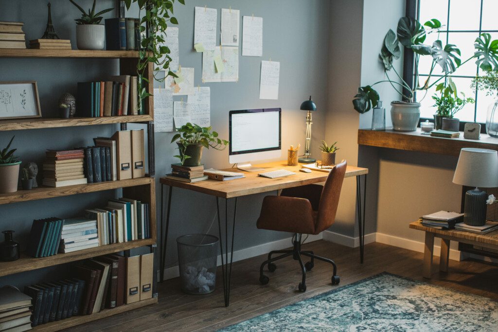 Escritório em casa com mesa de trabalho, computador, armário com livros e folhas coladas na parede. 