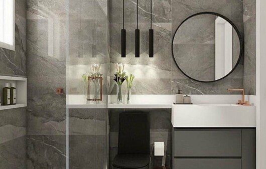 Banheiro cinza com revestimento em mármore, luminária pendente, espelho redondo e vaso preto.