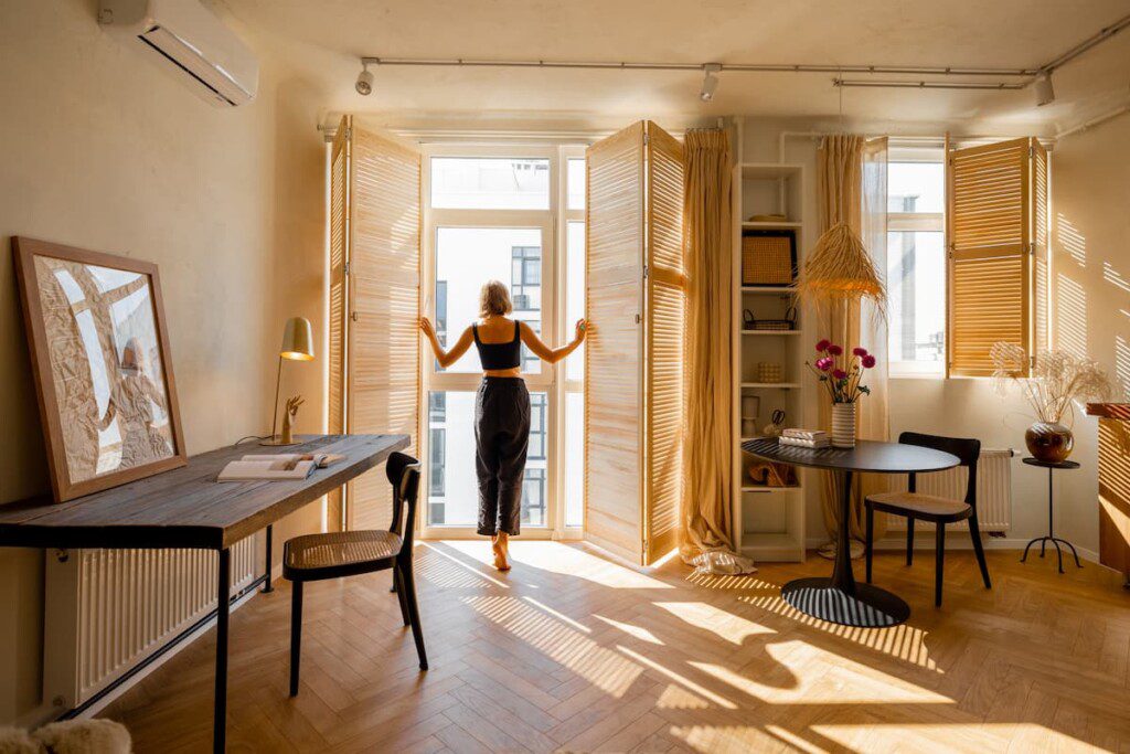 Mulher abre uma janela com persiana de madeira em uma quarto espaçoso e bem iluminado. 