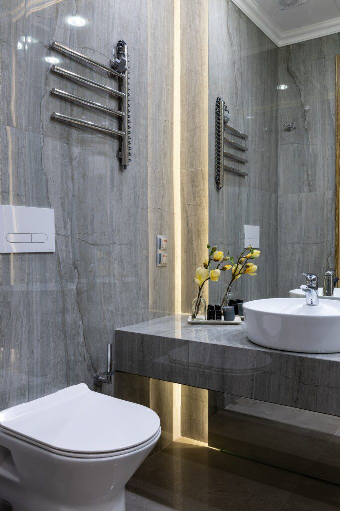 Banheiro com revestimento cinza que imita mármore, louças brancas e acessórios na cor preta. 