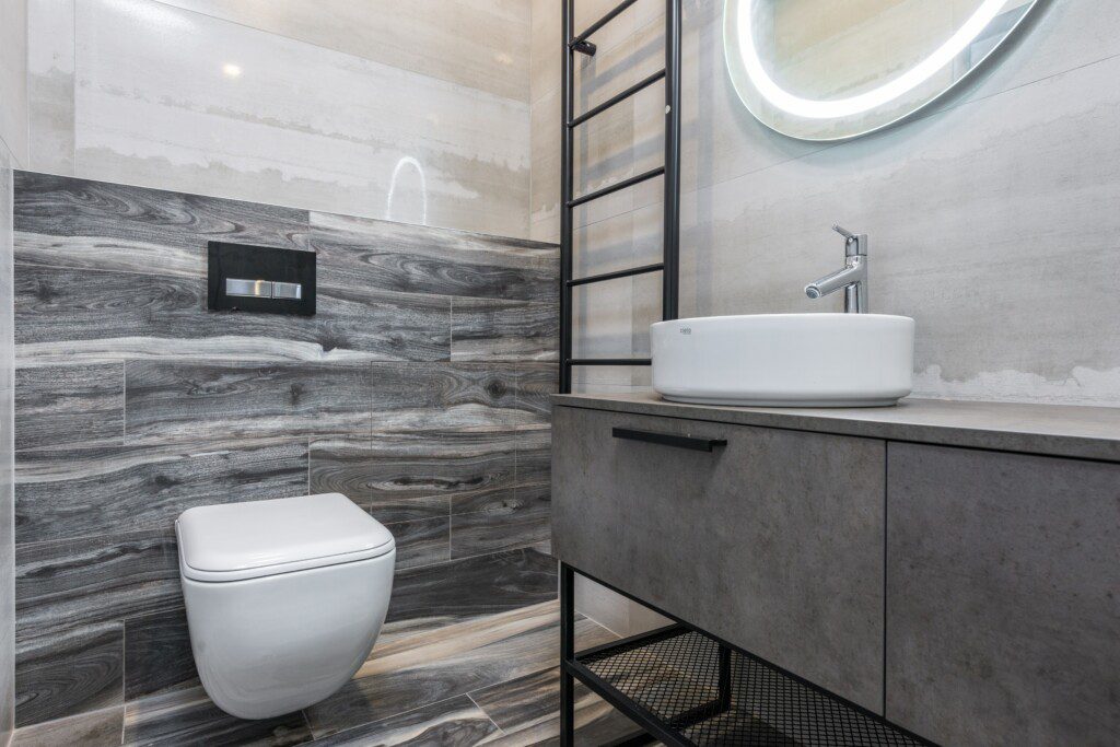 Banheiro com móveis e revestimento na cor cinza, espelho com luz de LED e acessórios em ferro preto. 
