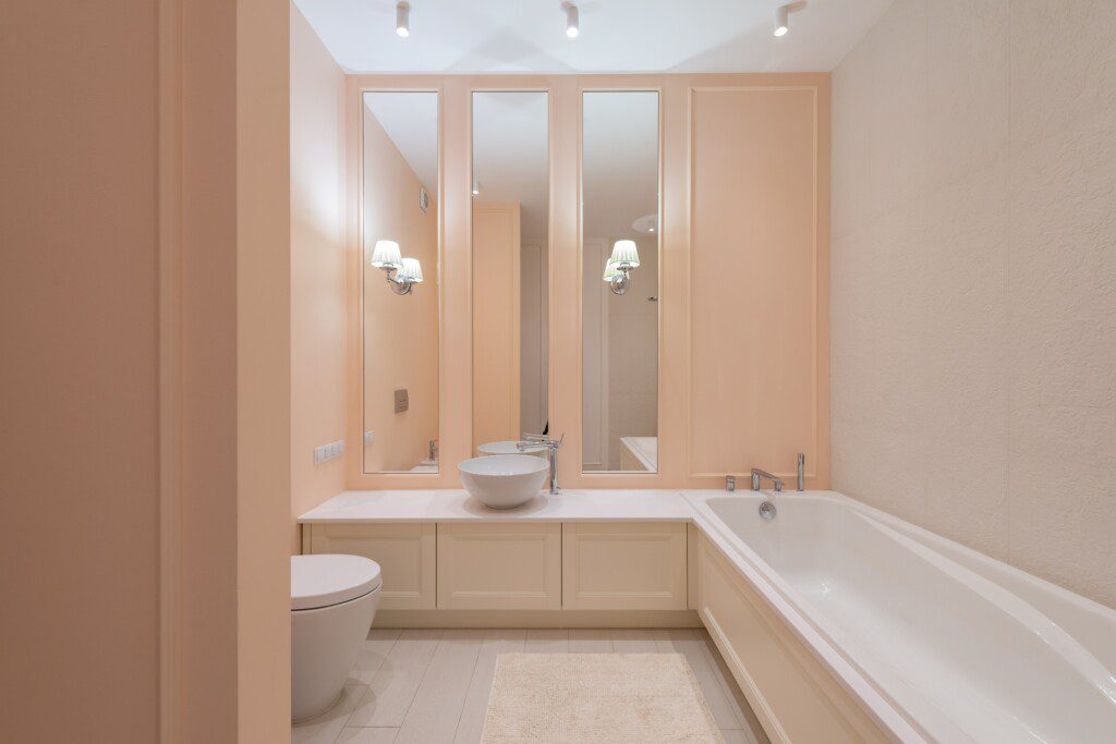 Banheiro bege com espelhos em três partes, banheira e detalhes em boiserie na parede. 