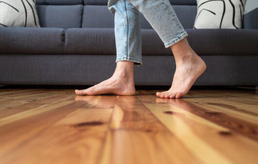 Uma mulher de pés descalços caminha sobre um piso de taco, é possível ver um sofá no fundo da foto.