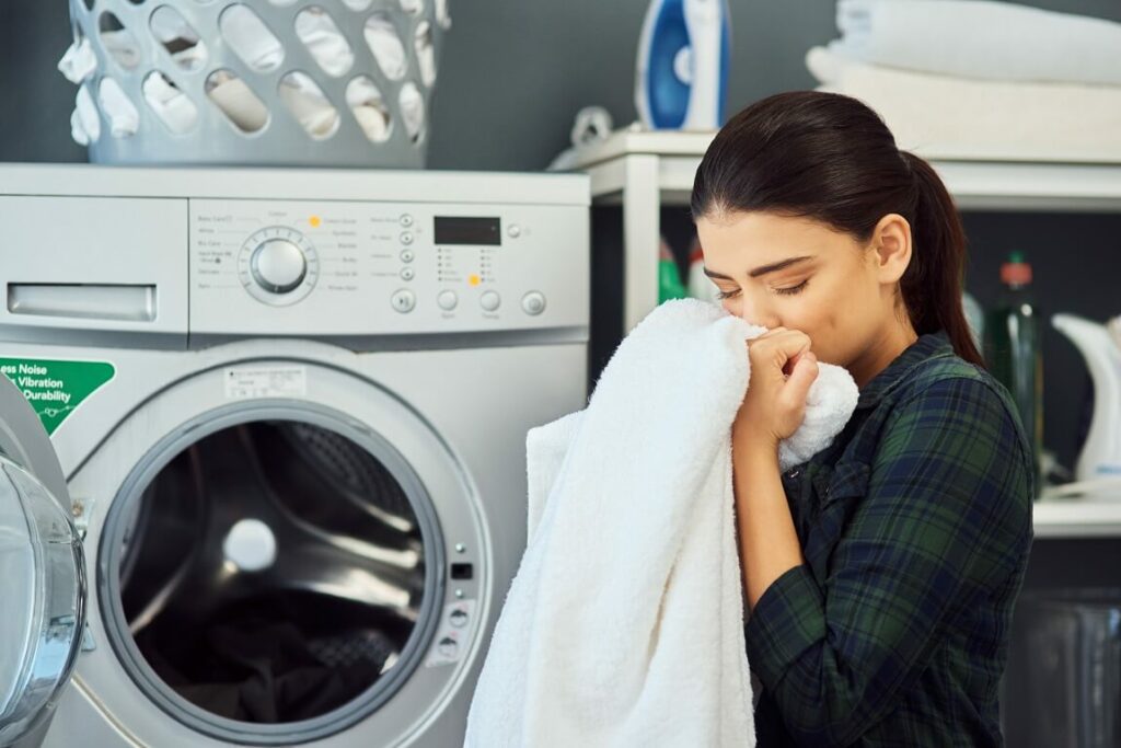 Foto que ilustra matéria com dicas sobre como deixar a casa cheirosa mostra uma mulher cheirando uma toalha branca diante de uma máquina de lavar roupas.