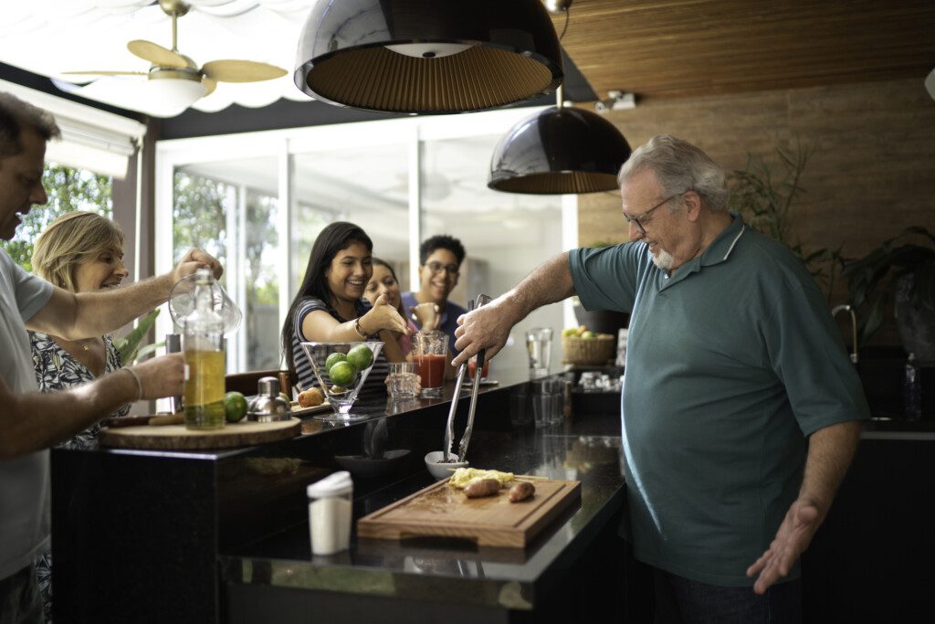 Imagem de um senhor manuseando ingredientes do churrasco atrás de um balcão com familiares reunidos do outro lado do cantinho do churrasco