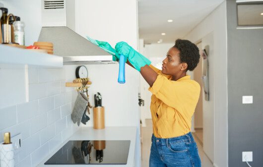magem de uma mulher usando luvas verdes descartáveis e segurando um produto de limpeza enquanto higieniza uma coifa de inox