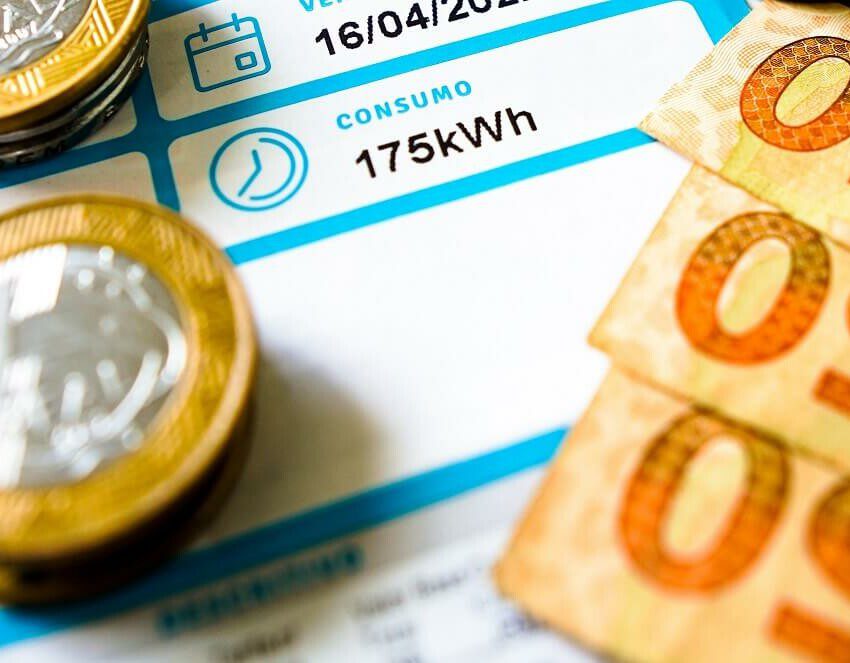 Foto que ilustra matéria sobre consumo de energia mostra uma conta de energia elétrica com moedas e notas em cima.