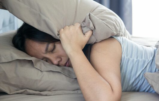 Foto que ilustra matéria sobre isolamento acústico mostra uma mulher deitada em uma cama de olhos fechados segurando um travesseiro sobre a cabeça