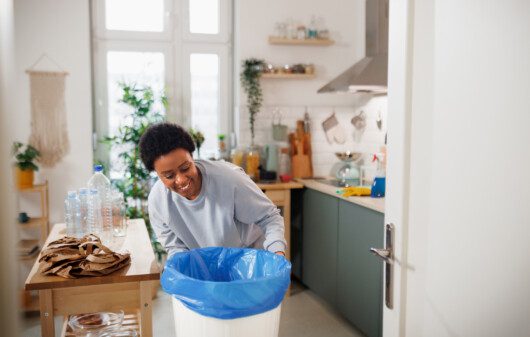 Imagem de uma mulher colocando um saco de lixo azul em uma lixeira branca na cozinha de casa