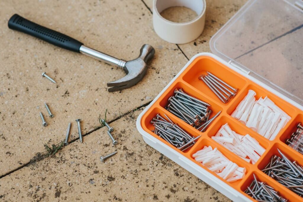 Foto que ilustra matéria sobre tipos de ferramentas mostra um martelo no chão ao lado de uma caixa repleta de parafusos.