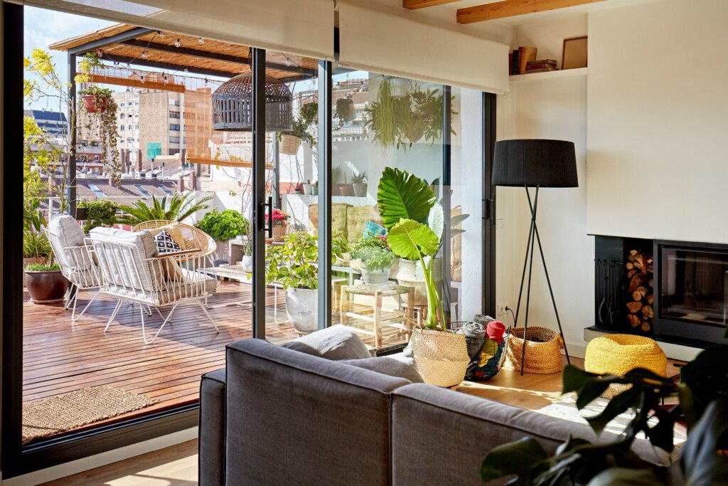 Imagem de parte da sala e uma varanda com piso de madeira, decorada com plantas, poltronas e uma mesa de centro
