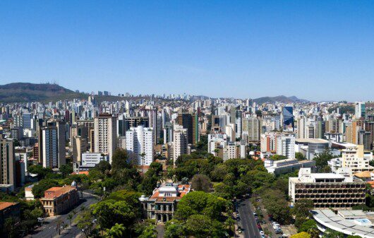 Foto que ilustra matéria sobre as regionais de Belo Horizonte mostra uma vista do alto da cidade, com destaque, me primeiro plano, para árvores da Praça da Liberdade. Ao fundo, prédios e mais atrás a Serra do Curral e um céu azul em um dia claro.