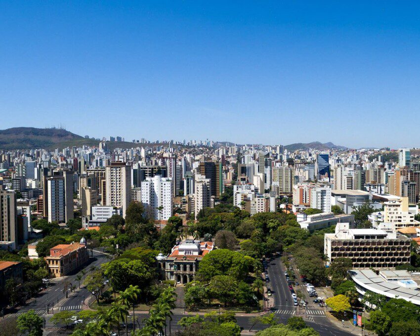 Foto que ilustra matéria sobre as regionais de Belo Horizonte mostra uma vista do alto da cidade, com destaque, me primeiro plano, para árvores da Praça da Liberdade. Ao fundo, prédios e mais atrás a Serra do Curral e um céu azul em um dia claro.
