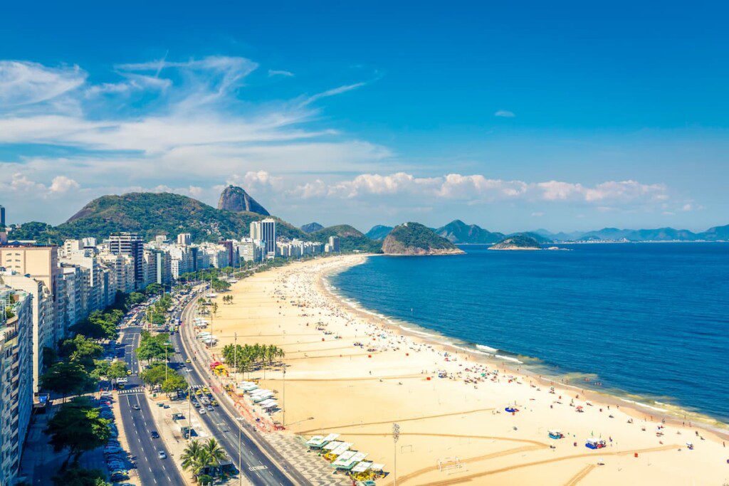 Imagem aérea da Praia de Copacabana. É possível ver o calçadão de Copacabana, prédios, faixa de areia, morros e o oceano.