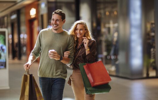 Imagem de um casal sorridente andando pelos corredores de um shopping com algumas sacolas de compras para ilustrar matéria sobre os shoppings no rj