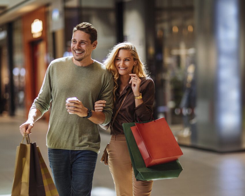 Imagem de um casal sorridente andando pelos corredores de um shopping com algumas sacolas de compras para ilustrar matéria sobre os shoppings no rj