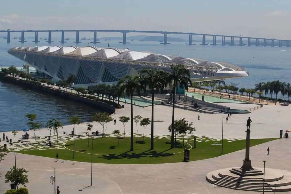 Foto que ilustra matéria sobre a Zona Portuária do Rio de Janeiro mostra o Museu do Amanhã visto do alto, com a Baía de Guanabara e a Ponte Rio-Niterói em um dia de sol.