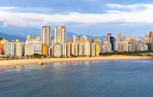 Foto que ilustra matéria sobre o que fazer em São Vicente mostra uma vista panorâmica da faixa de areia da praia de Itararé, com o mar em primeiro plano e prédios altos ao fundo