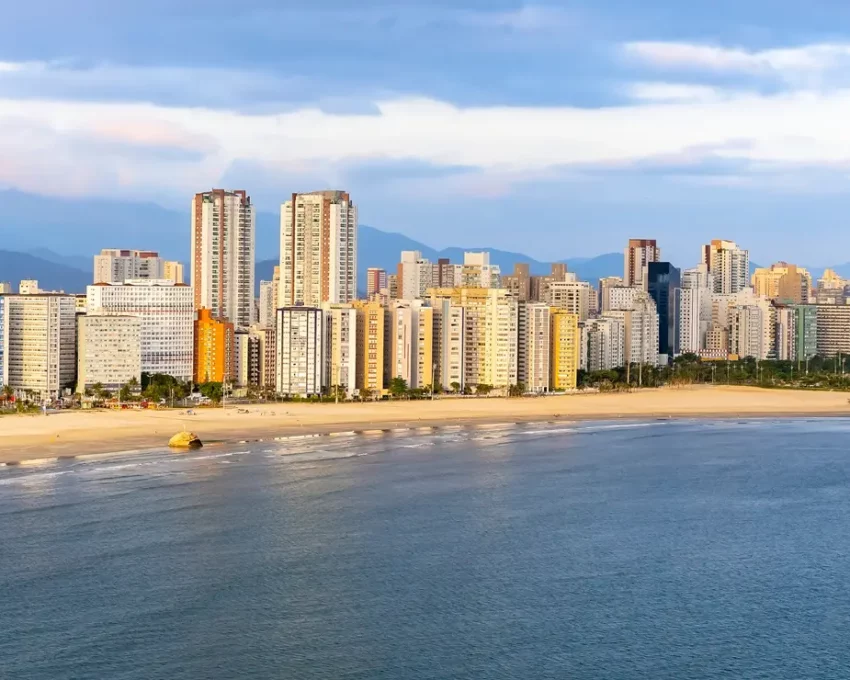 Foto que ilustra matéria sobre o que fazer em São Vicente mostra uma vista panorâmica da faixa de areia da praia de Itararé, com o mar em primeiro plano e prédios altos ao fundo