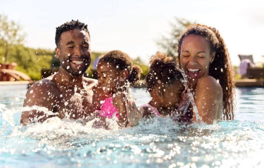 Imagem de uma família em uma piscina com duas meninas espirrando água para cima para ilustrar matéria sobre clubes em SP