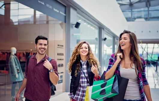 Imagem de três amigos, sendo duas mulheres e um homem, passeando sorridentes e com sacolas nas mãos pelo corredor de um shopping para ilustrar matéria sobre shopping em Goiânia