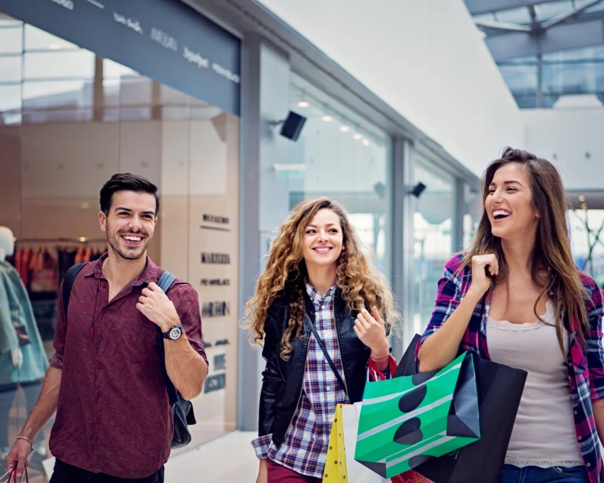 Imagem de três amigos, sendo duas mulheres e um homem, passeando sorridentes e com sacolas nas mãos pelo corredor de um shopping para ilustrar matéria sobre shopping em Goiânia