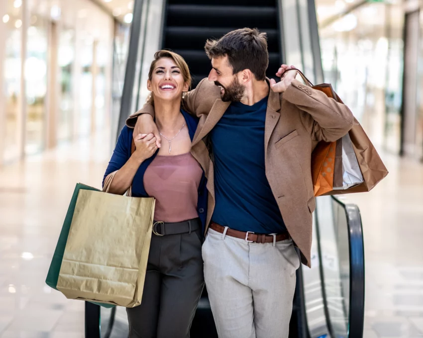 Imagem de um homem e uma mulher abraçados e felizes, descendo uma escada rolante com sacolas de compras nas mãos, para ilustrar matéria sobre shopping em Guarulhos