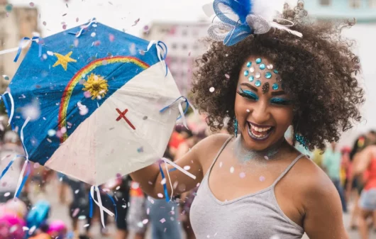 Foto que ilustra matéria sobre Carnaval de Recife mostra uma mulher negra, muito sorridente e enfeitada com glitter e brilhantes diversos se divertindo com uma pequena sombrinha usada para dançar frevo com estampa da bandeira do estado de Pernambuco. (Foto: Getty Images)