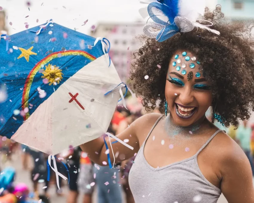 Foto que ilustra matéria sobre Carnaval de Recife mostra uma mulher negra, muito sorridente e enfeitada com glitter e brilhantes diversos se divertindo com uma pequena sombrinha usada para dançar frevo com estampa da bandeira do estado de Pernambuco. (Foto: Getty Images)
