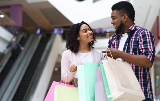 Imagem de um casal negro, composto por um homem e uma mulher, felizes com sacolas de compras nas mãos para ilustrar matéria sobre os melhores shoppings de SP