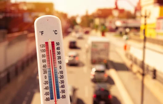 Fotografia de uma avenida muito iluminada pelo sol. Uma pessoa segura um termômetro que marca alta temperatura.
