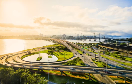 Imagem aérea de carros nos viadutos e pistas próximos ao mar durante o nascer do sol em Florianópolis para ilustrar matéria sobre cidades tranquilas para morar