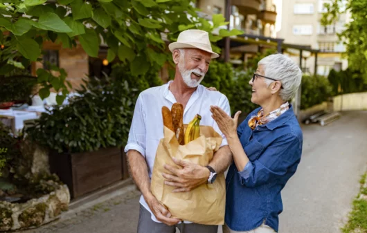 Imagem de um casal de idosos segurando uma sacola de papel com pães e caminhando pela rua, com um prédio e árvores atrás, para ilustrar matéria sobre as melhores cidades para aposentados morarem no Brasil