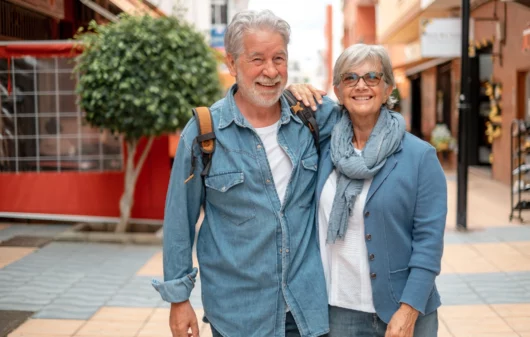 Imagem de um casal de idosos composto por um homem e uma mulher brancos, de cabelos brancos, sorridentes e vestindo camisetas jeans para ilustrar matéria sobre cidade para idosos no Brasil
