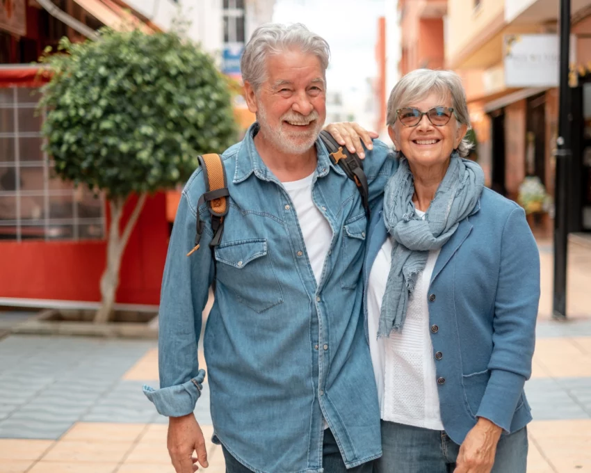 Imagem de um casal de idosos composto por um homem e uma mulher brancos, de cabelos brancos, sorridentes e vestindo camisetas jeans para ilustrar matéria sobre cidade para idosos no Brasil