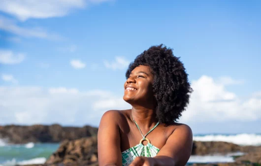 Imagem de uma mulher negra sorridente vestindo uma camisa azul sentada em uma praia olhando para o lado com um céu azul ao fundo para ilustrar matéria sobre as cidades mais seguras da Bahia