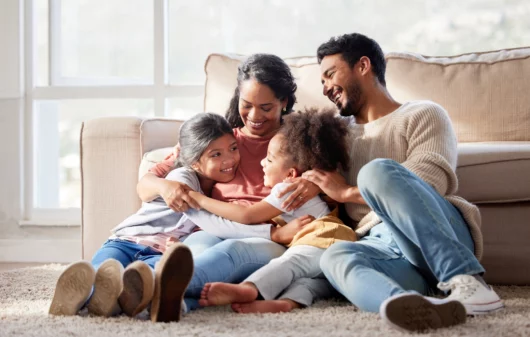 Imagem de uma família composta por um homem, uma mulher e duas crianças felizes no chão da sala para ilustrar matéria sobre o estado mais seguro do Brasil