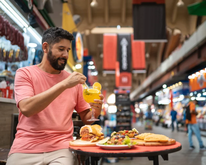 Imagem de um homem tomando uma bebida com alguns lanches em cima de uma mesa no Mercado Municipal para ilustrar matéria sobre os mercados em São Paulo