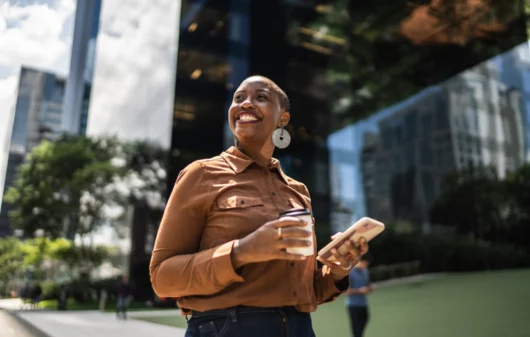 Imagem de uma mulher negra andando em uma rua em frente a um prédio empresarial, sorrindo e com um celular na mão, para ilustrar matéria sobre qual a cidade mais tranquila do Brasil