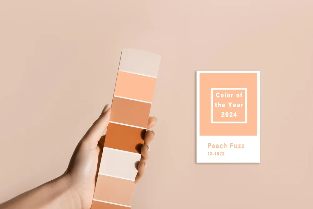 Foto que ilustra matéria sobre a cor do ano 2024 da Pantone mostra uma mão segurando uma paleta de cores. Ao lado, há um card em inglês com as menções a "Color of the year", à cor Peach Fuzz e a seu código na tabela Pantone, que é 13-1023 (Foto: Shutterstock)