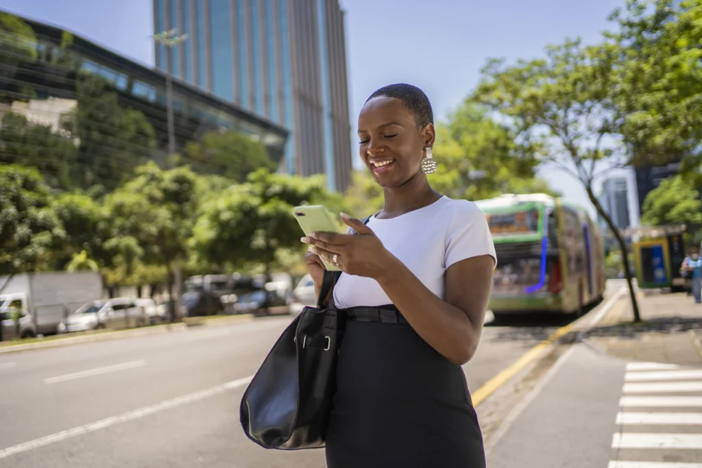 Imagem de uma mulher negra de cabelo curto com um celular na mão e sorriso no rosto em um distrito financeiro de São Paulo para ilustrar matéria sobre como é morar em Santos e trabalhar em São Paulo