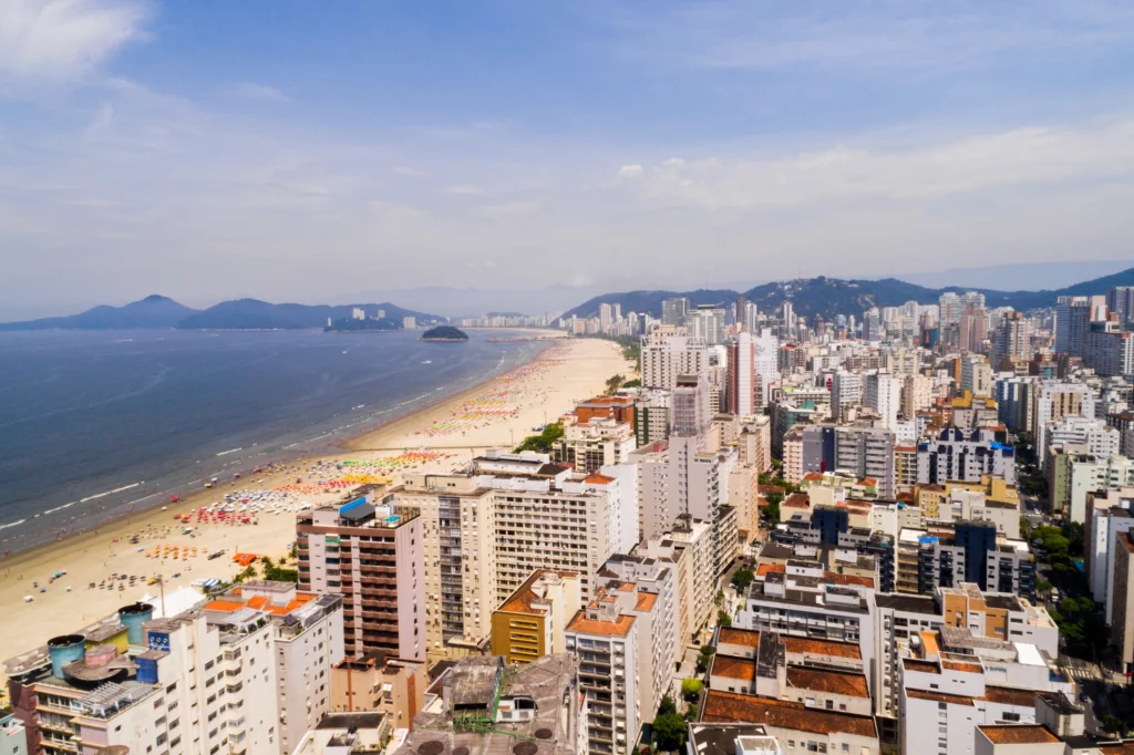 Vista aérea da paisagem urbana de Santos mostra mar, faixa de areia e prédios à beira-mar para ilustrar matérias sobre morar em Santos e trabalhar em São Paulo