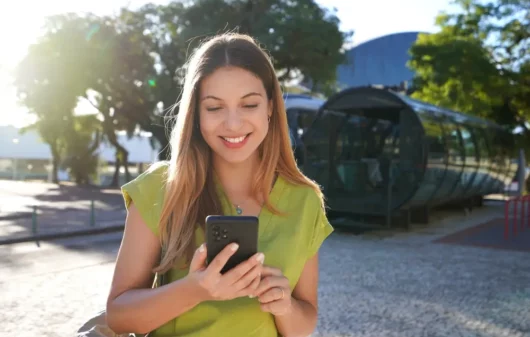 Foto que ilustra matéria sobre Cidades inteligentes no Brasil mostra uma mulher sorrindo enquanto olha para um celular próxima a uma estação de ônibus de Curitiba (Foto: Shutterstock)